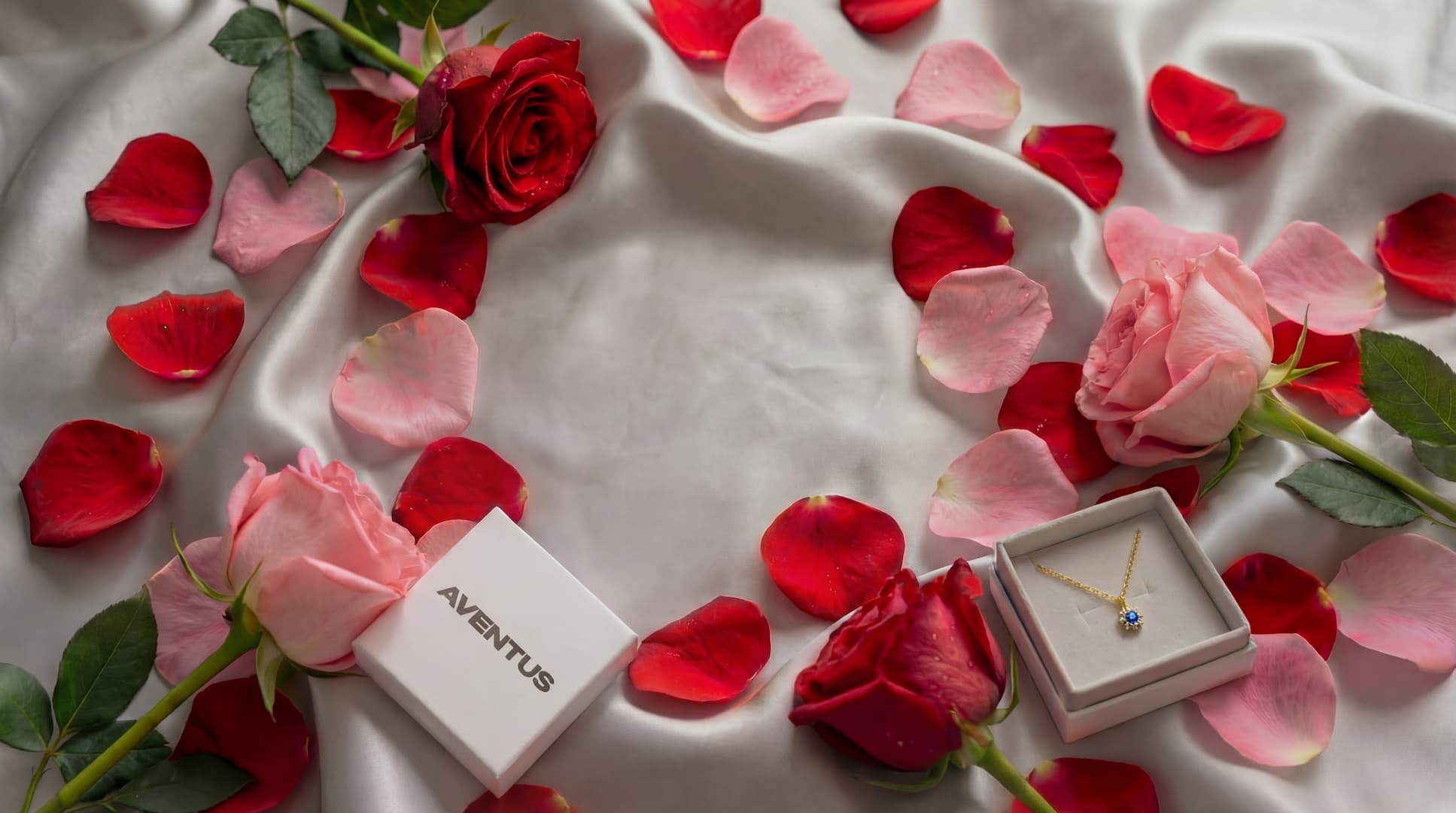 Diamond necklace in a box surrounded by red and pink roses and petals on a white fabric background